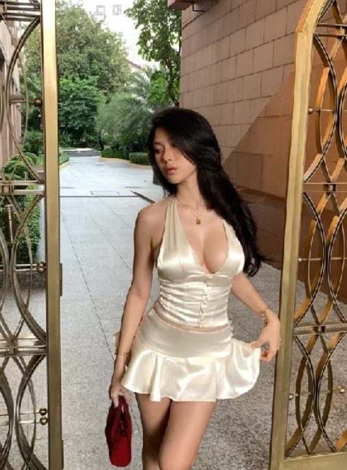 A young woman with long black hair poses outdoors in a satin halter top and matching mini skirt, holding a red handbag.