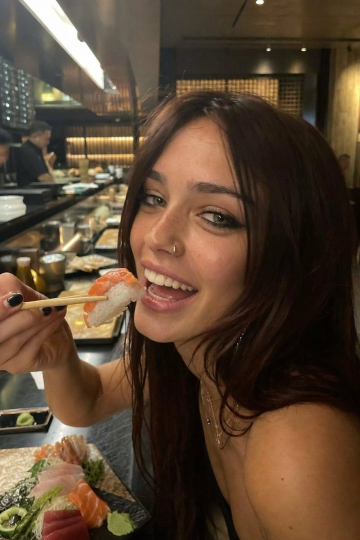 A young woman with long brown hair and green eyes smiles as she eats sushi with chopsticks in a restaurant.