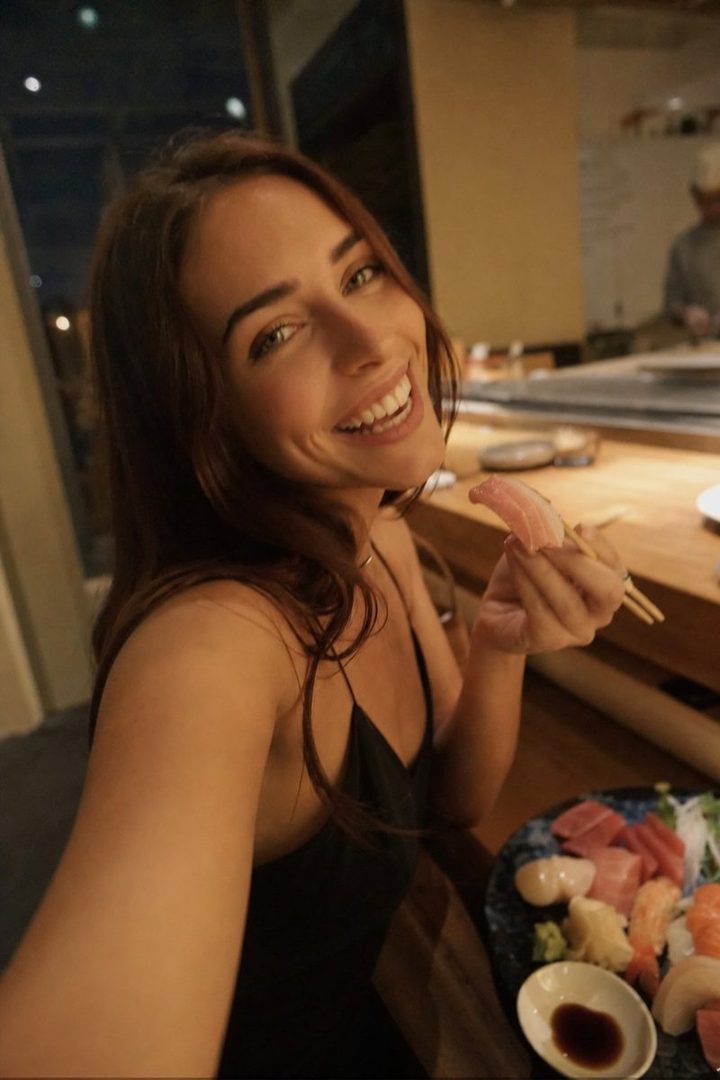 A smiling woman with long brown hair takes a selfie while eating sushi at a restaurant.