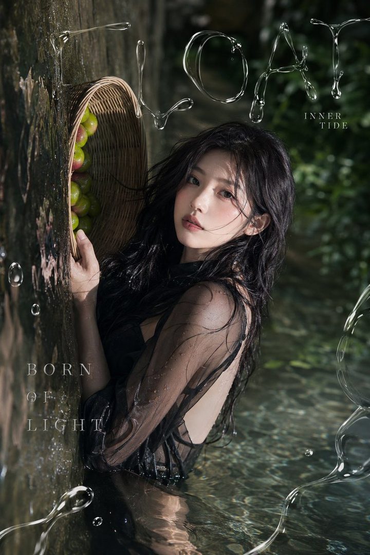 A young woman with long black hair, wearing a sheer black dress, sits in shallow water surrounded by nature, with a basket of apples nearby. The image has a dreamy, ethereal quality.