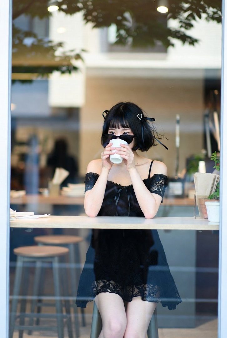 A young woman with short black hair and heart-shaped hair clips, wearing sunglasses and a black off-the-shoulder dress, drinks from a white cup while sitting at a cafe table.