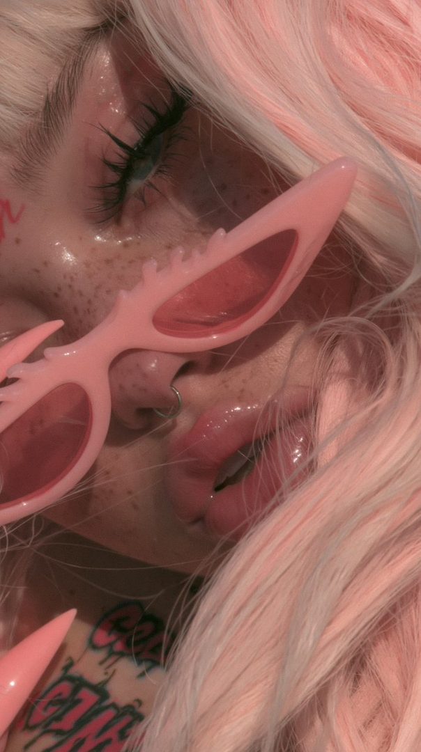 Close-up portrait of a young woman with long pink hair, wearing unique pink sunglasses and showing off her makeup and tattoos.