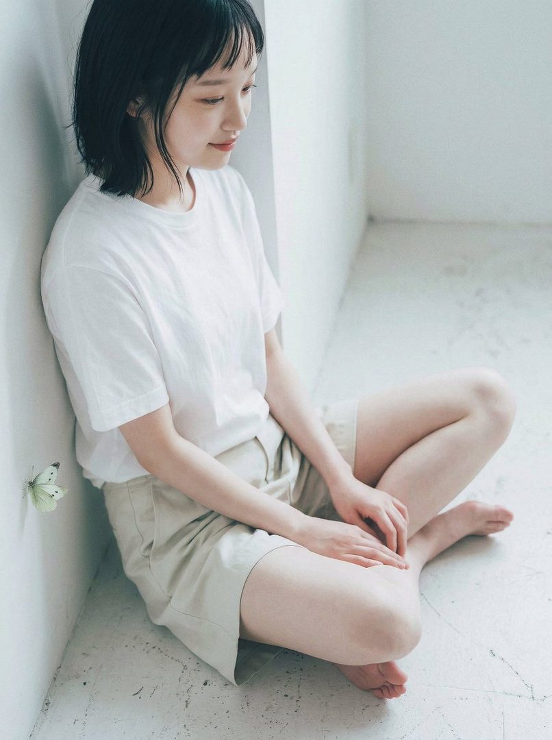A young woman with short black hair sits cross-legged indoors, wearing a white t-shirt and beige shorts, with a butterfly on the wall.