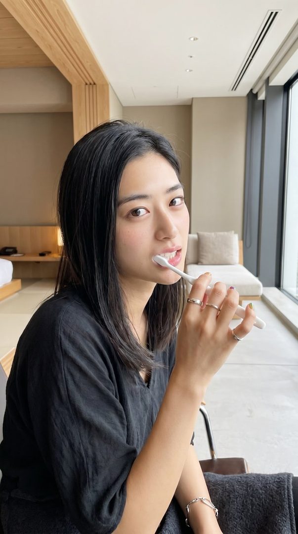 A young East Asian woman with long black hair brushing her teeth in a hotel room.