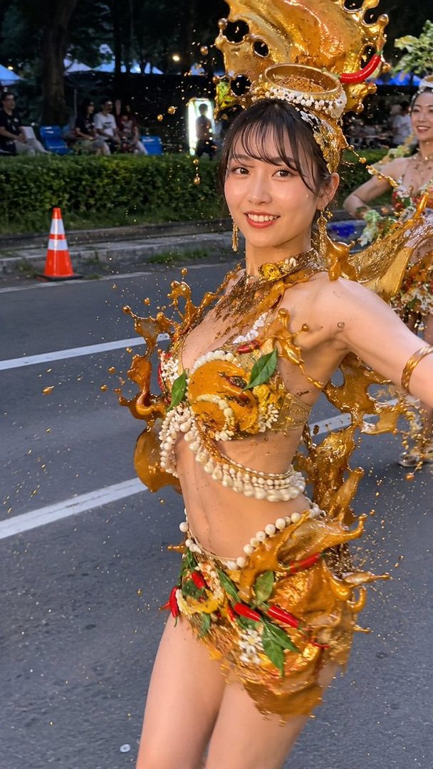 A smiling young woman in a golden, elaborate costume with splashes of liquid, possibly during a parade or festival.