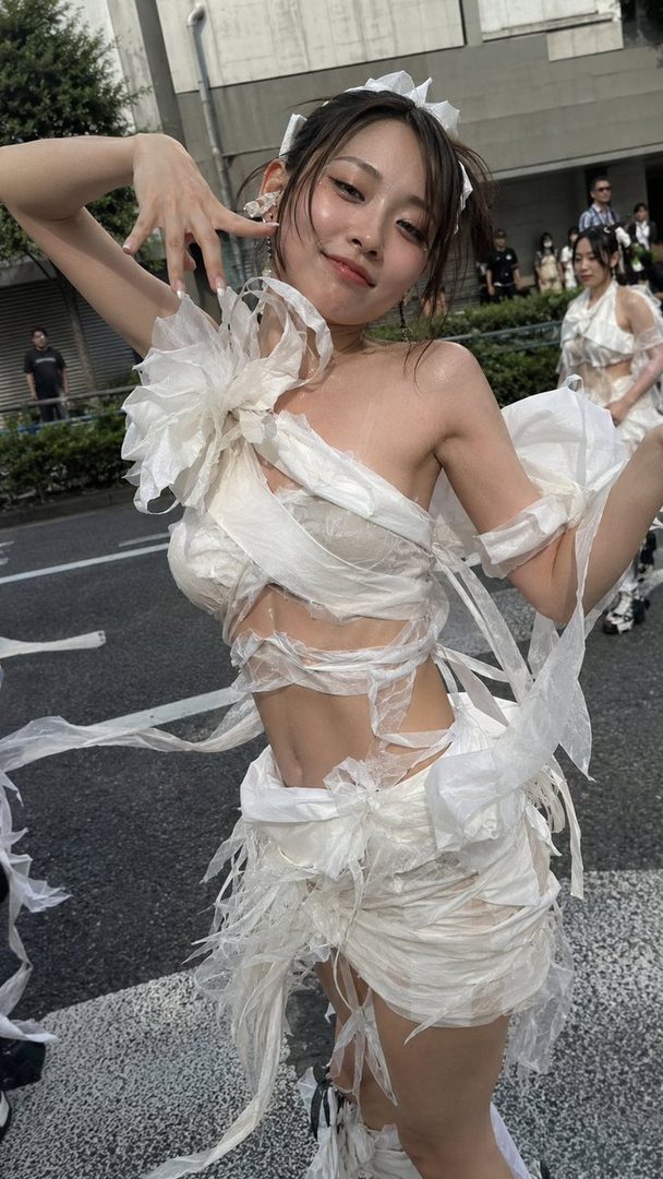 A Japanese woman in a unique white tissue-like outfit poses playfully on a street during a fashion show.