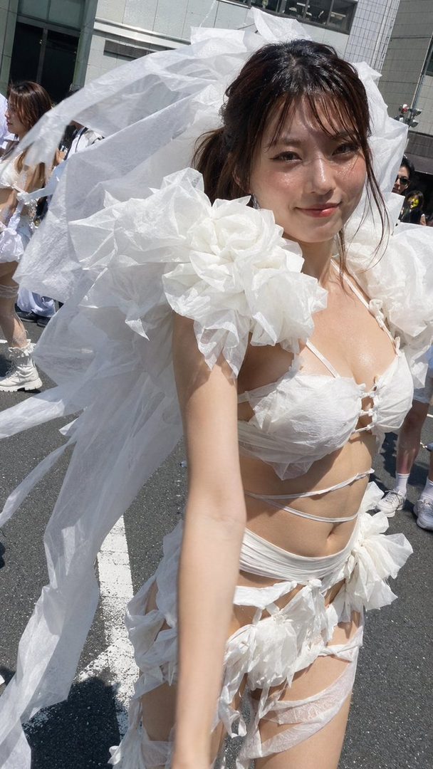 A young Japanese woman in a creative white outfit resembling tissue paper, posing outdoors on a street during a fashion event.