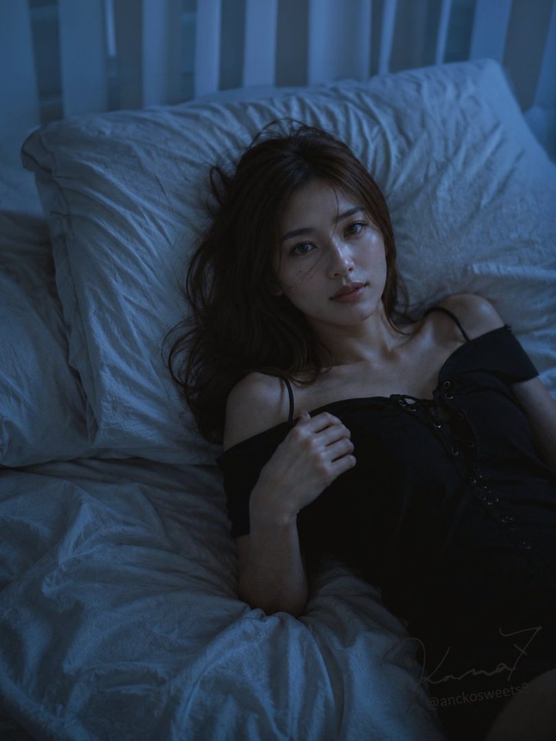 A young woman with long brown hair lies on a bed in a black off-the-shoulder dress, looking seductively at the camera.