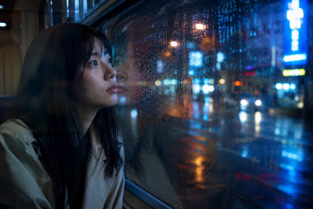 A young woman with long black hair looks out a window on a rainy night in the city, with neon lights reflecting on the wet street.