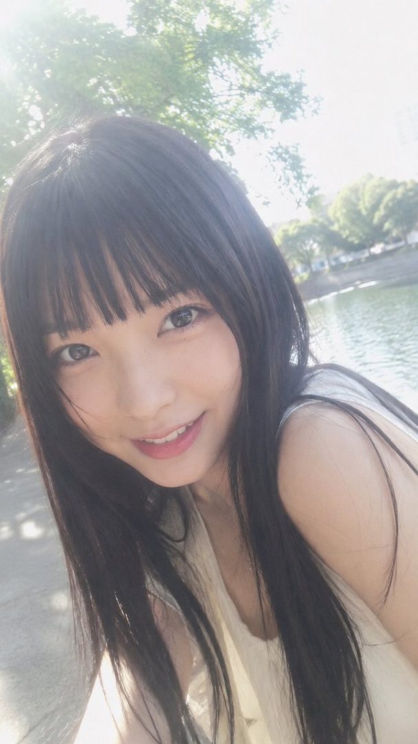 A young East Asian woman with long black hair and bangs smiles at the camera in a close-up selfie taken outdoors.
