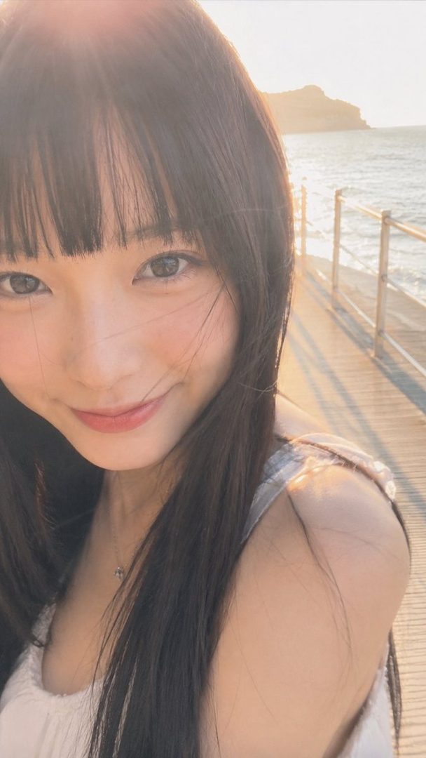 A young woman with long dark hair and bangs smiles gently at the camera, standing on a wooden pier by the sea during golden hour.