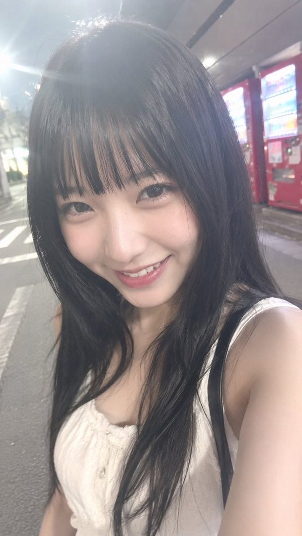 A young East Asian woman with long black hair and bangs smiles at the camera in a selfie taken outdoors at night, with vending machines in the background.