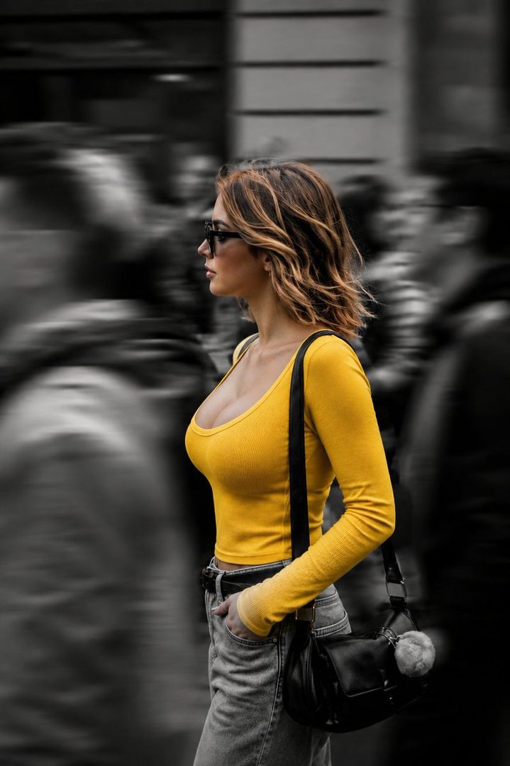 A woman in a yellow long-sleeved top and jeans walks down a blurred street, wearing sunglasses and carrying a black handbag.
