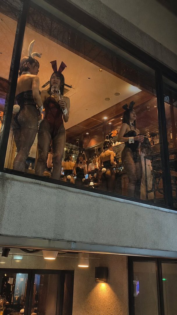Several women in bunny costumes posing in a bar, viewed through a window.