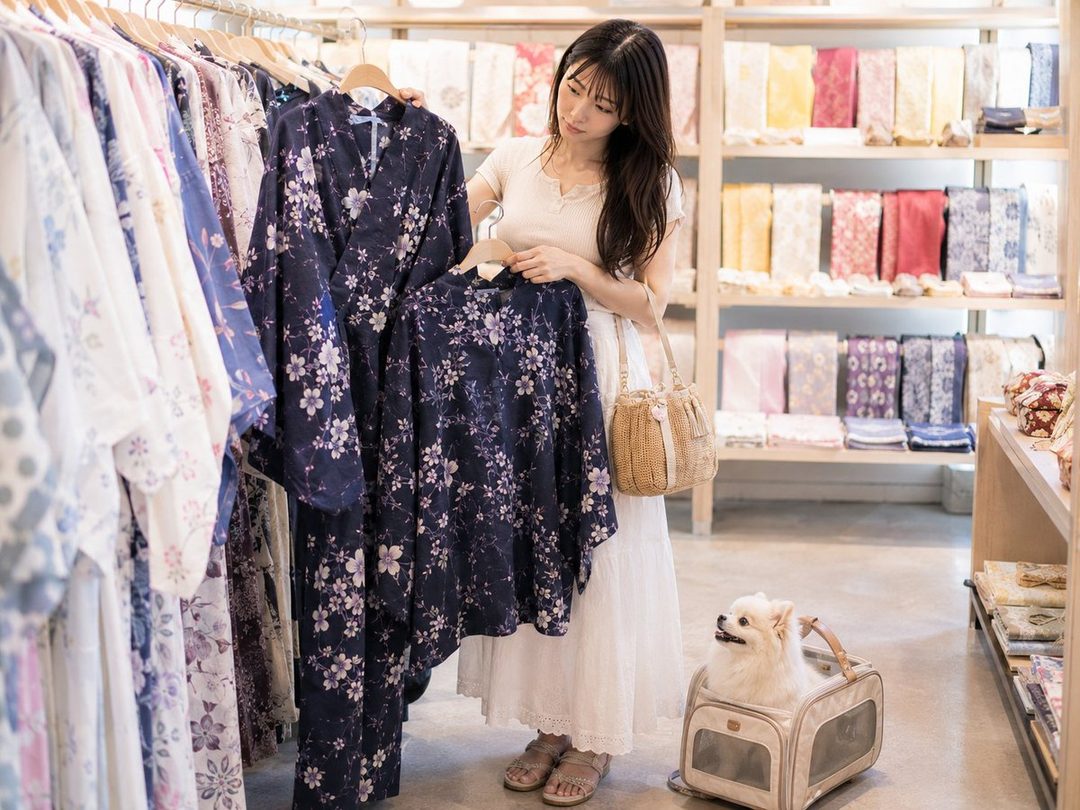 A young woman with long dark hair is browsing yukatas in a shop, holding two floral patterned yukatas, with a small white dog in a carrier at her feet.