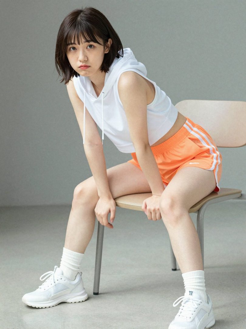 A young woman with short black hair, wearing a white sleeveless hoodie and orange athletic shorts, sits on a chair in a studio setting.