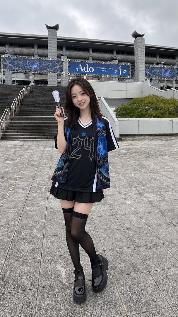 A young woman in a black skirt and thigh-high stockings stands in front of a stadium, holding a light stick.