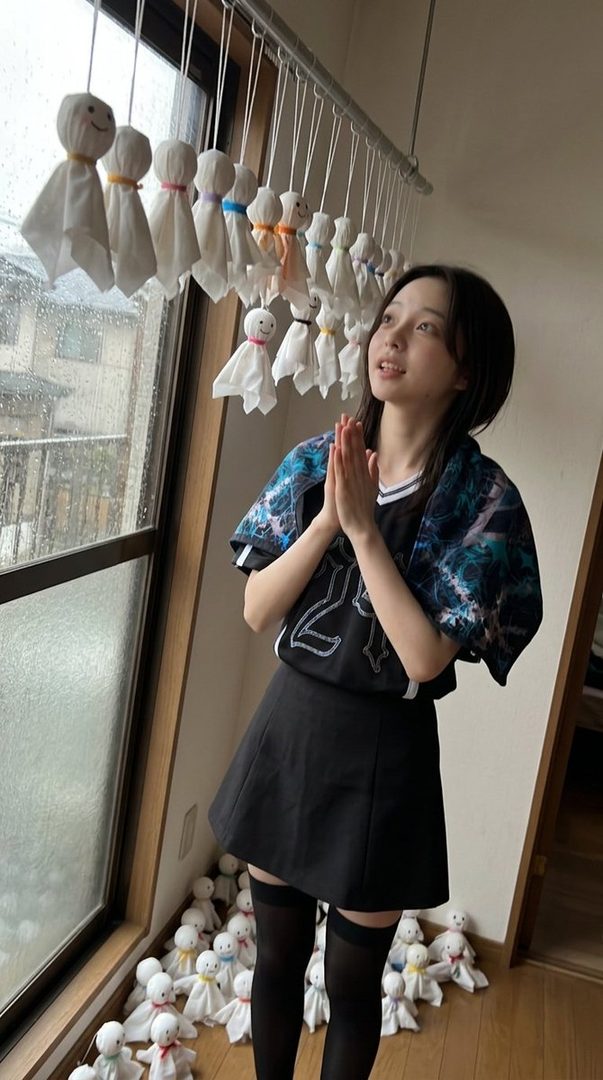 A young Japanese woman with short black hair stands indoors, wearing a black skirt and thigh-high stockings, with her hands clasped in prayer. She is looking up towards a row of traditional Japanese rain dolls (teru teru bozu) hanging from a rack.