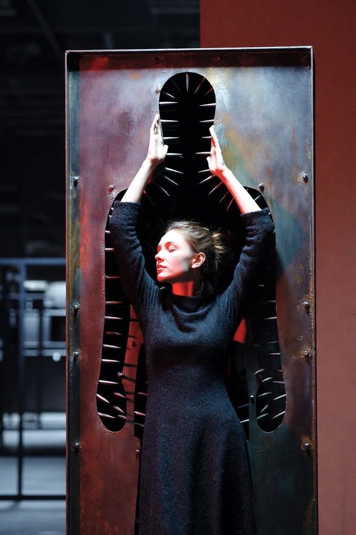 A woman in a dark dress stands with her arms raised inside a metal frame with a silhouette cutout.