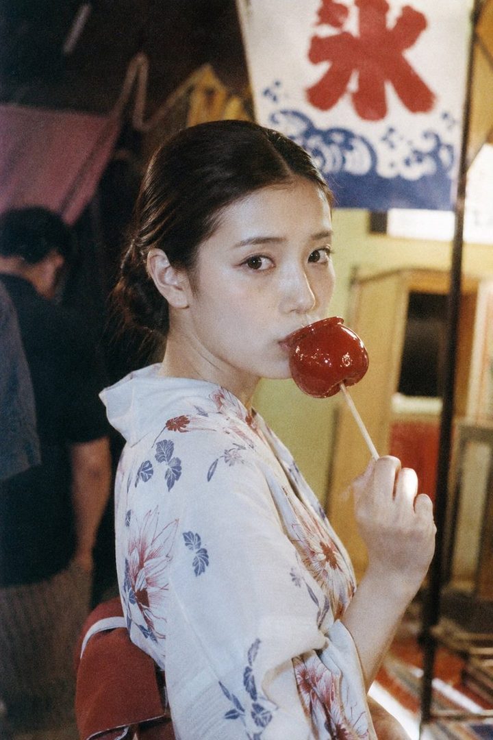 A young Japanese woman in a floral kimono eats a candied apple at a summer festival.