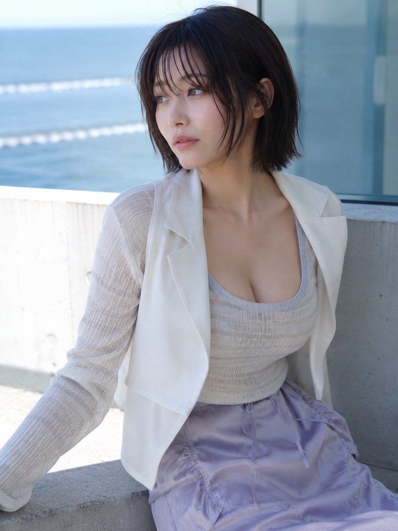 A young woman with short dark hair and bangs, wearing a light cardigan over a tank top and a lavender skirt, looking thoughtfully towards the ocean.