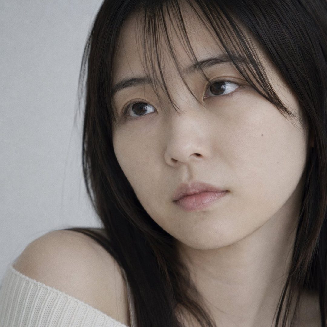 Close-up portrait of a young Japanese woman with dark hair, wearing a cream off-shoulder knit sweater, looking slightly upwards with a dreamy expression.