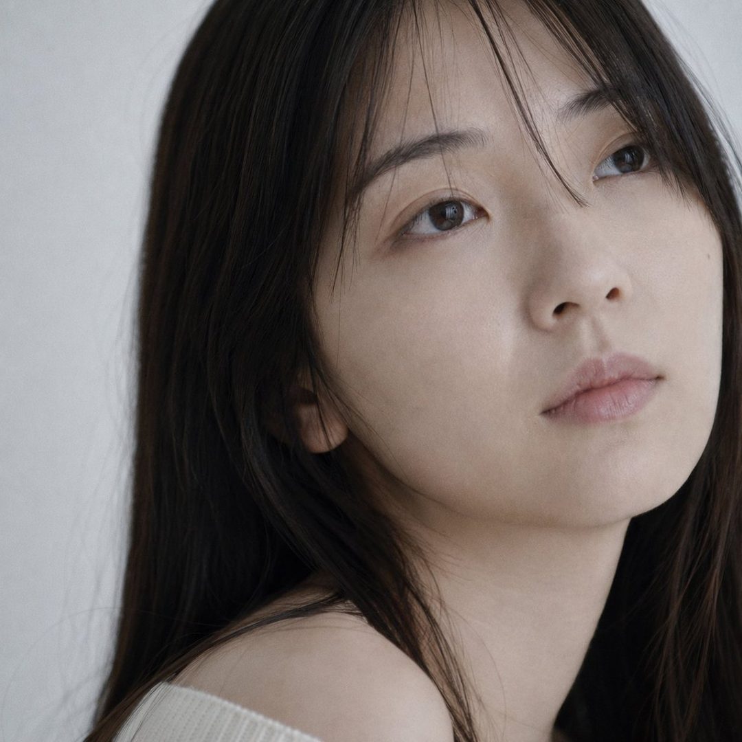 Close-up portrait of a young Japanese woman with long dark hair, looking upwards with a soft expression, wearing an off-shoulder cream knit sweater.