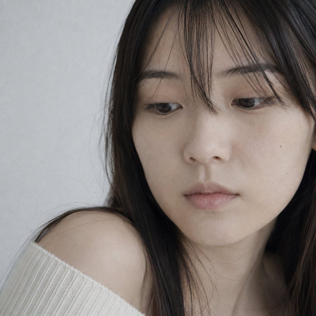 Close-up portrait of a young Japanese woman with long dark hair, wearing a cream knit sweater, looking down with a dreamy expression.