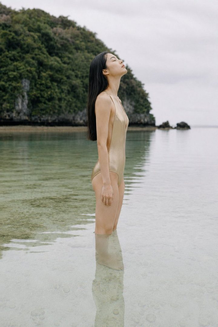 A woman in a gold one-piece swimsuit stands in shallow, clear water, with her eyes closed and head tilted back, facing a lush green island in the background.
