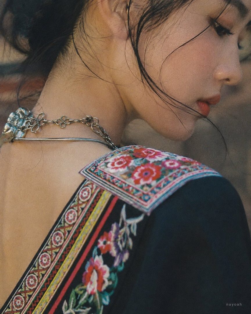 Close-up portrait of a young East Asian woman with dark hair, wearing a black blouse with colorful floral embroidery and a silver necklace.