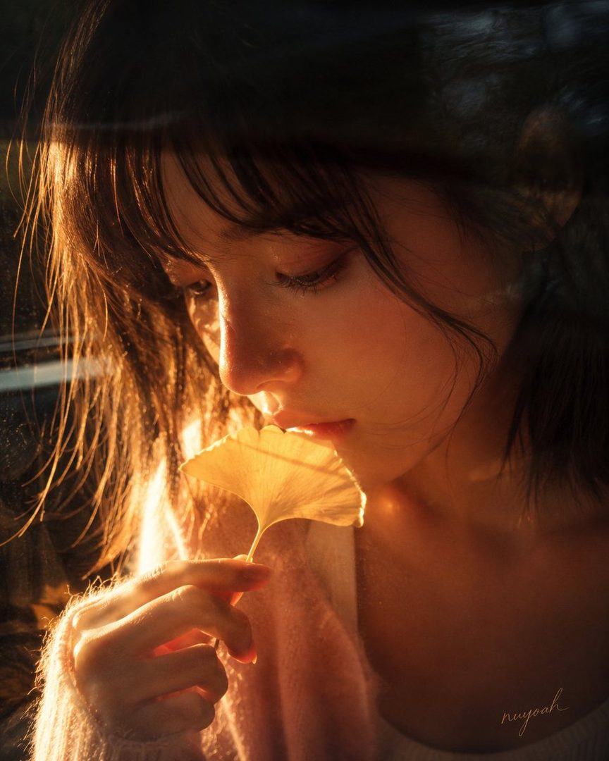 A young woman with dark hair holds a golden ginkgo leaf in the warm glow of golden hour light.