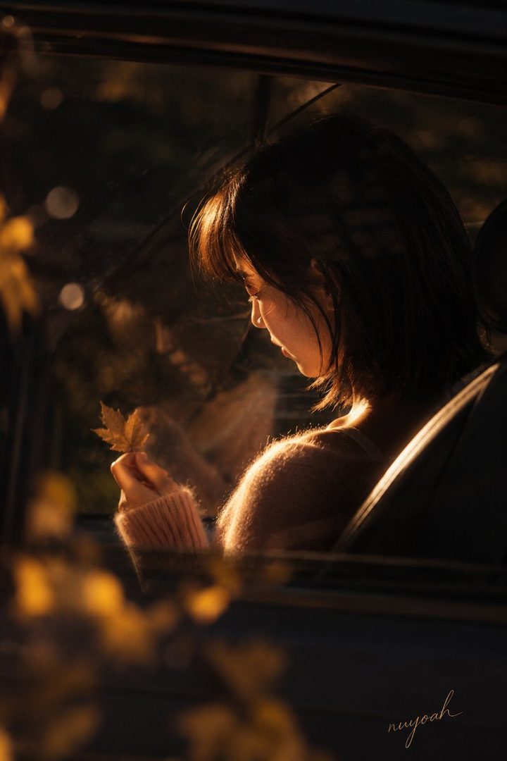 A young woman sits in a car, holding a fallen leaf, illuminated by the warm, golden light of sunset filtering through the car window.
