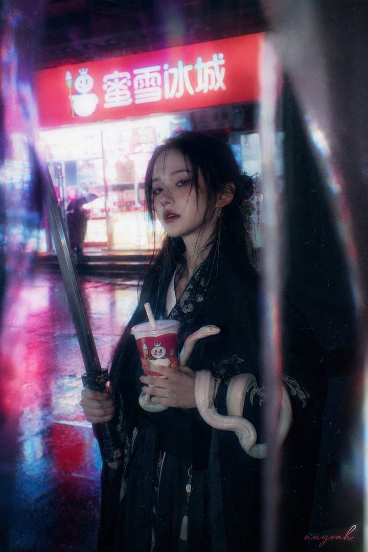 A young woman with long black hair, dressed in traditional dark clothing, holds a sword and a drink cup while a snake coils around her arm, standing on a wet street at night with neon lights.