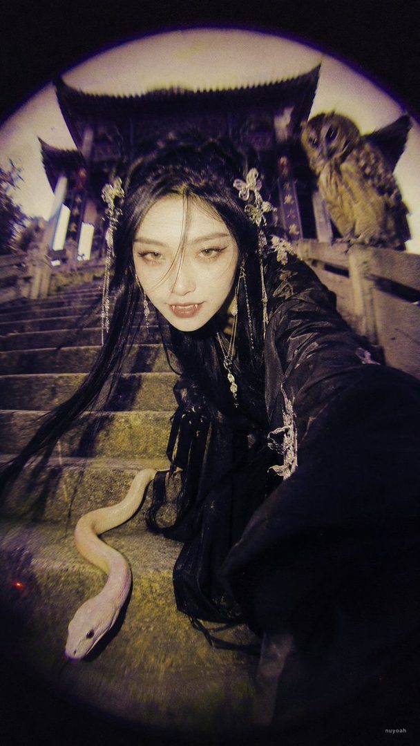 A woman with fangs and dark hair, dressed in traditional Chinese attire, poses with a snake and an owl on a stone staircase, evoking a gothic vampire aesthetic.