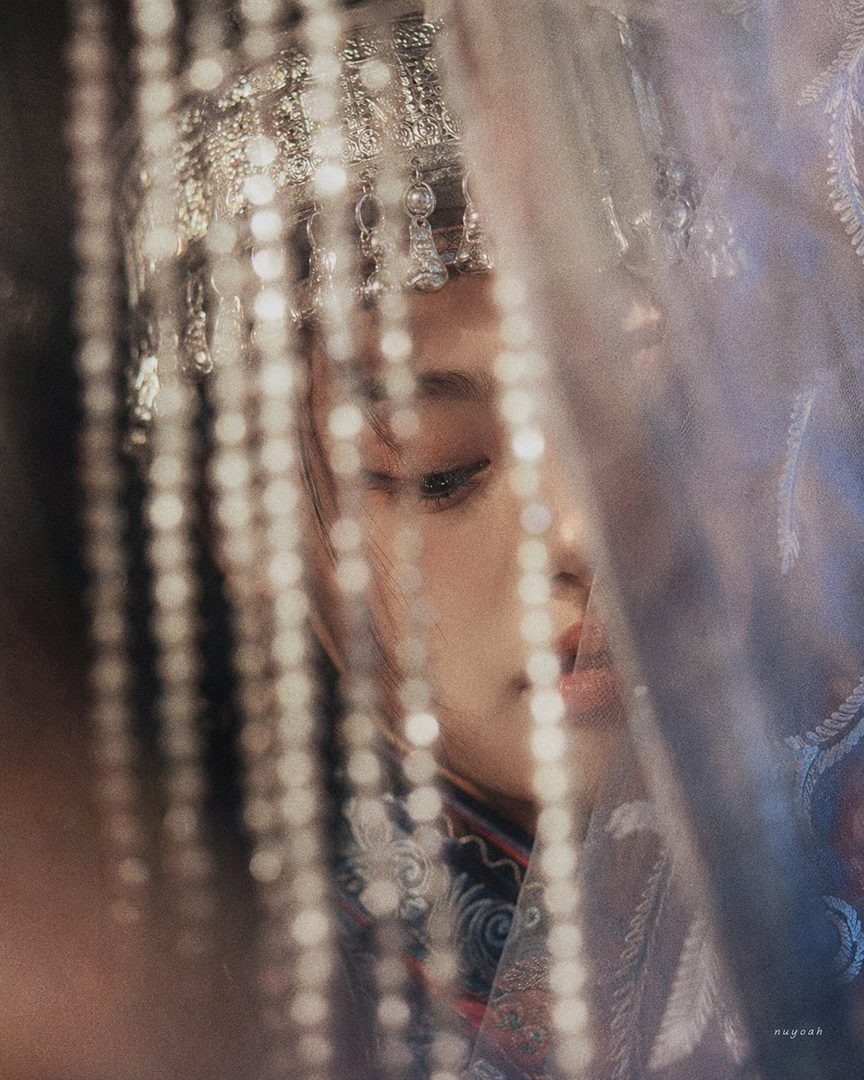 A close-up portrait of an East Asian woman wearing a traditional silver headdress and ornate clothing, partially obscured by sheer fabric and beaded curtains, with a dreamy and elegant mood.