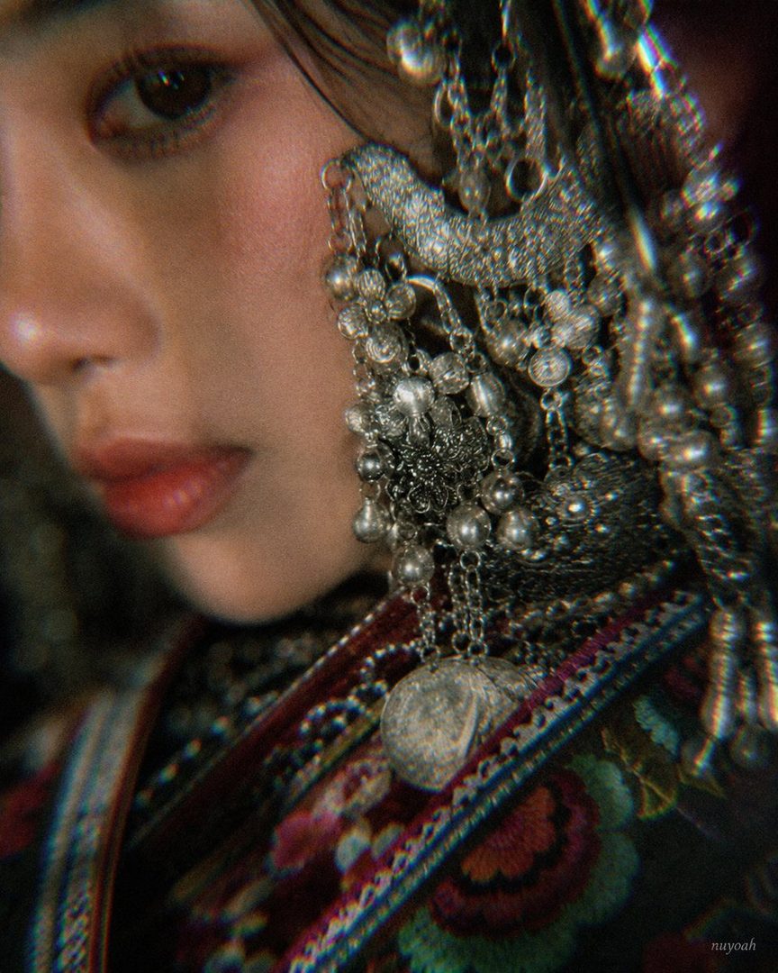 Close-up portrait of an East Asian woman with intricate silver jewelry and traditional embroidered clothing, her face partially obscured.