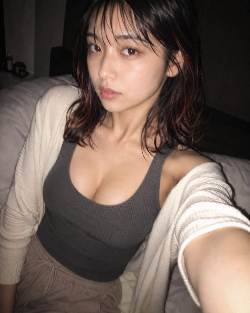 A young woman with wet, dark brown hair and red highlights, wearing a grey tank top and cream cardigan, takes a selfie in a dimly lit room.