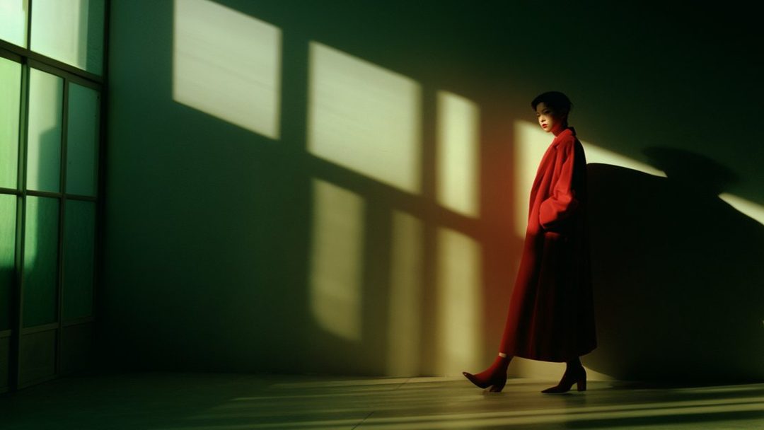 A woman in a red coat and boots stands in a room with dramatic shadows cast by a window.