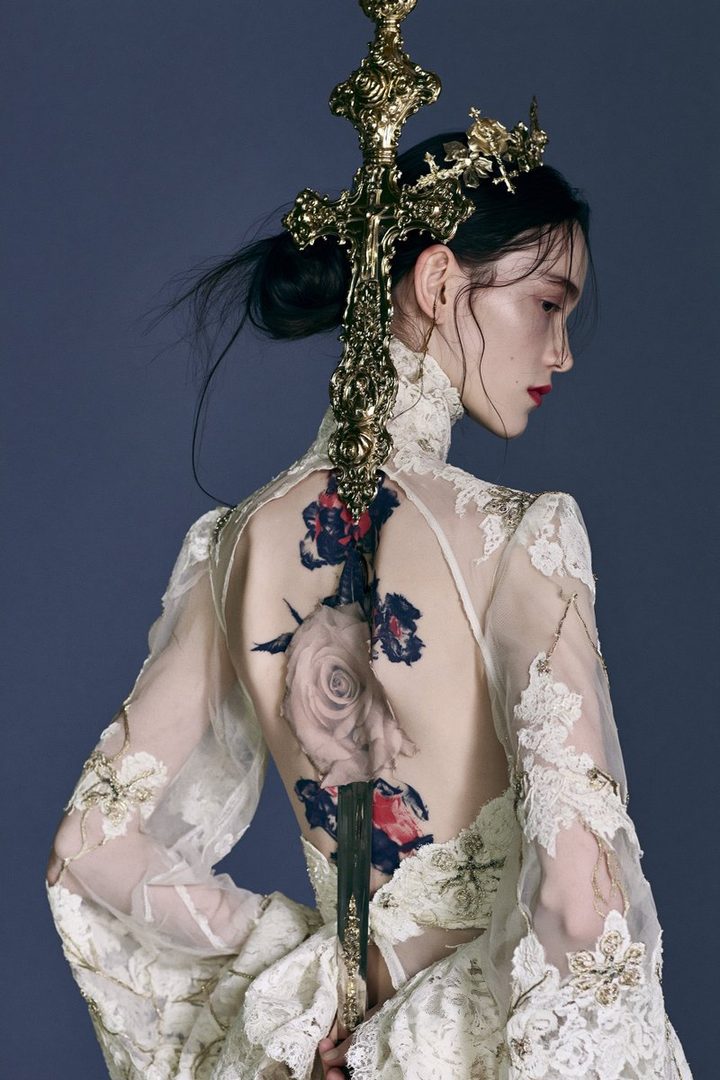 A woman with dark hair styled up, wearing an ornate white lace dress and a golden crown, holding a decorative golden cross staff, with a large rose tattoo on her back.