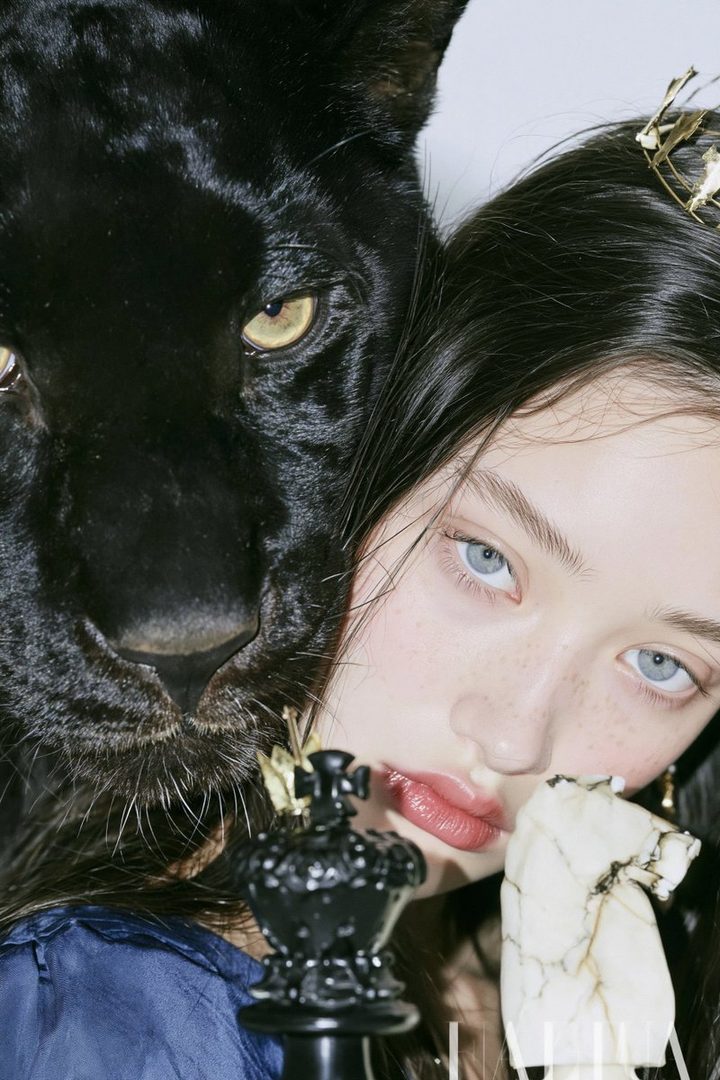 A young woman with striking blue eyes and freckles poses next to a black panther, holding a chess piece.