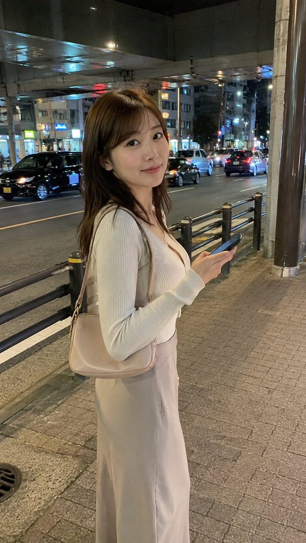 A young East Asian woman with long brown hair stands on a city street at night, holding a smartphone and looking at the camera with a gentle smile. She is wearing a light-colored ribbed top and a long skirt, with a beige shoulder bag.