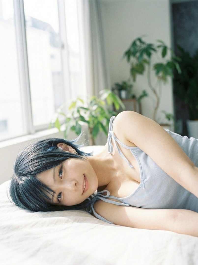 A young woman with short black hair lies on a bed, looking at the camera with a gentle smile. She is wearing a light blue camisole and is indoors near a window with plants.
