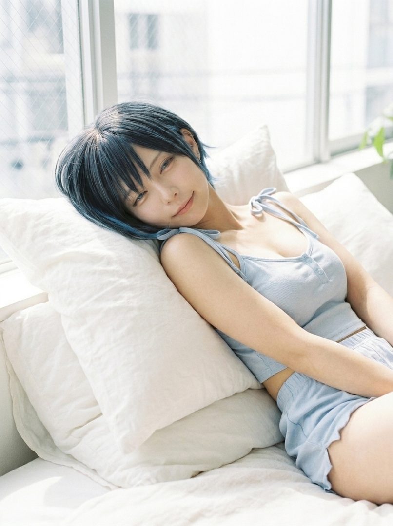 A young woman with short, dark hair with blue highlights, wearing a light blue camisole and shorts, rests on a bed with pillows, looking relaxed.