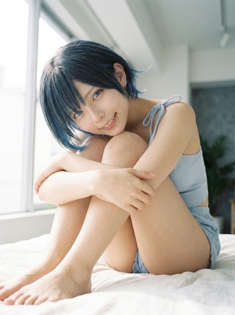 A young woman with short blue-black hair and bright eyes smiles gently while sitting on a bed, hugging her knees.