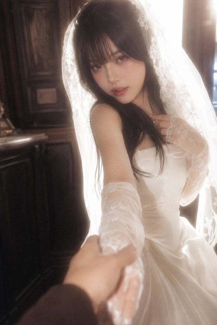 A young woman in a white wedding dress and veil, holding someone's hand, looking towards the camera with a dreamy expression.