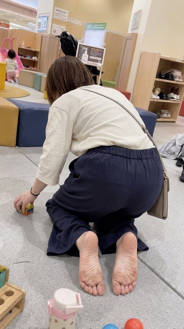 A woman in casual clothes kneels on the floor playing with a child in an indoor play area, showing the soles of her feet.