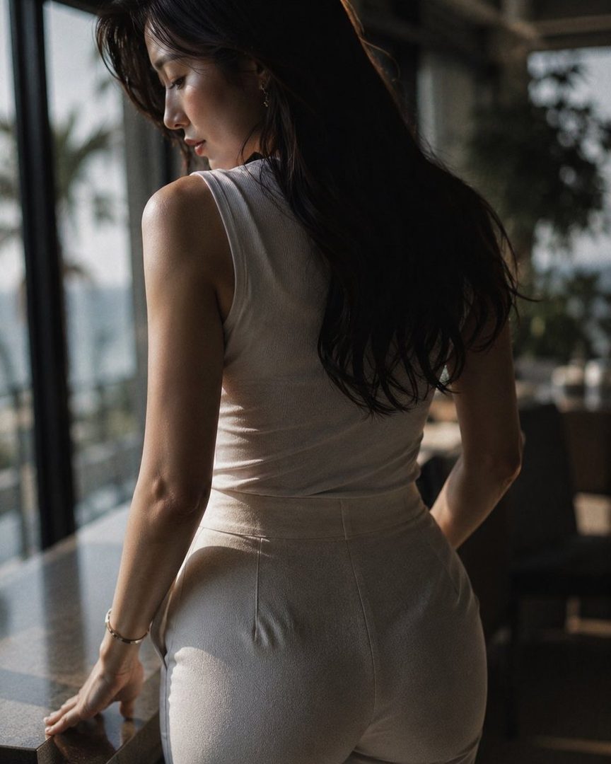 A woman with long dark hair, wearing a beige sleeveless top and pants, stands by a window looking down.