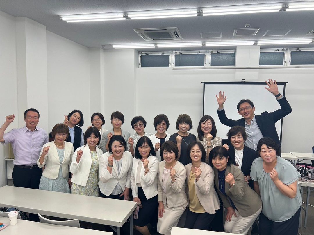 A group of 16 adults, mostly women, pose for a photo in an indoor meeting room, with some raising their fists in the air and one man waving.