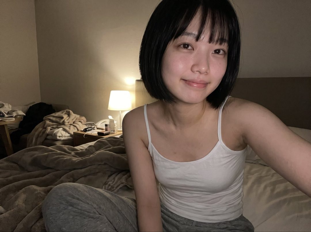 A young East Asian woman with short black hair and bangs, wearing a white tank top and grey sweatpants, smiles at the camera while sitting on a bed in a dimly lit bedroom.