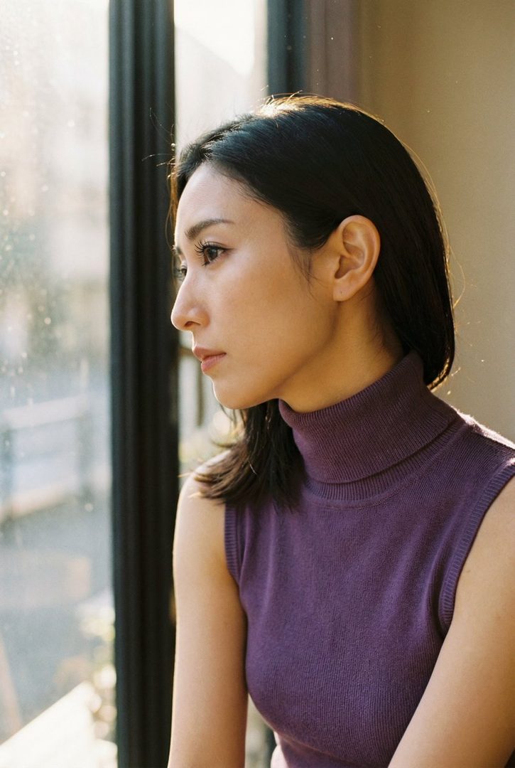 A woman with dark hair, wearing a purple sleeveless turtleneck, looks out a window.